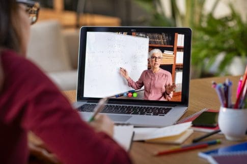 Student taking notes while looking at computer screen following professor doing math on video call.