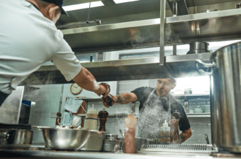 Two workers in food preparation jobs help each other by passing a pepper grinder