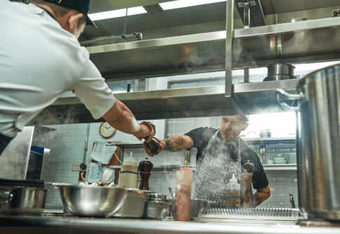 Two workers in food preparation jobs help each other by passing a pepper grinder